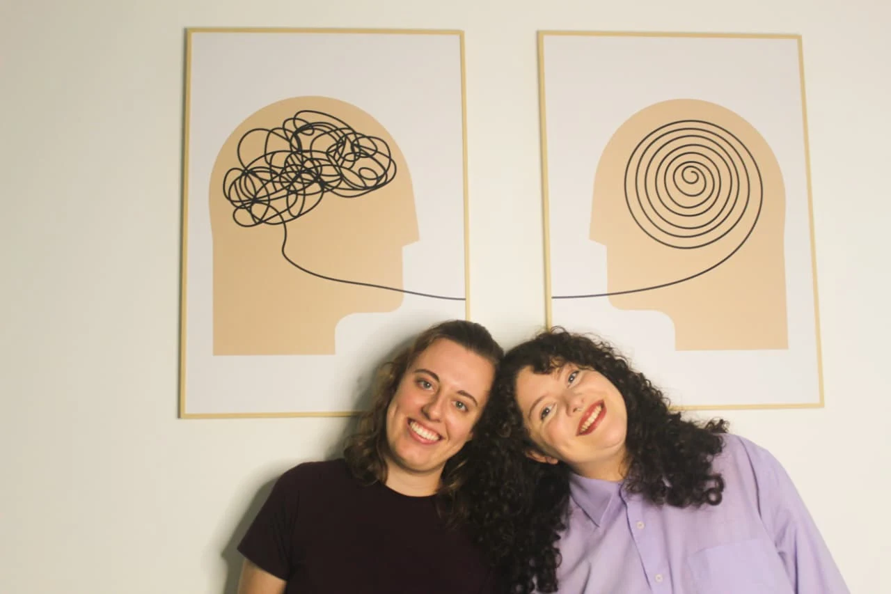 Ana Paula e Ana Clara sorrindo, encostadas uma na outra, sob dois quadros: uma cabeça com linhas emaranhadas e outra com uma espiral organizada